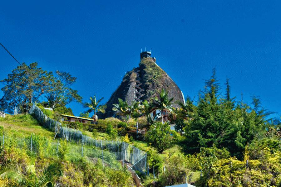 Un imposant monolithe rocheux pointu, équipé d'un escalier en zigzag dans sa faille, domine une colline verdoyante parsemée de végétation et de maisons sous un ciel bleu.