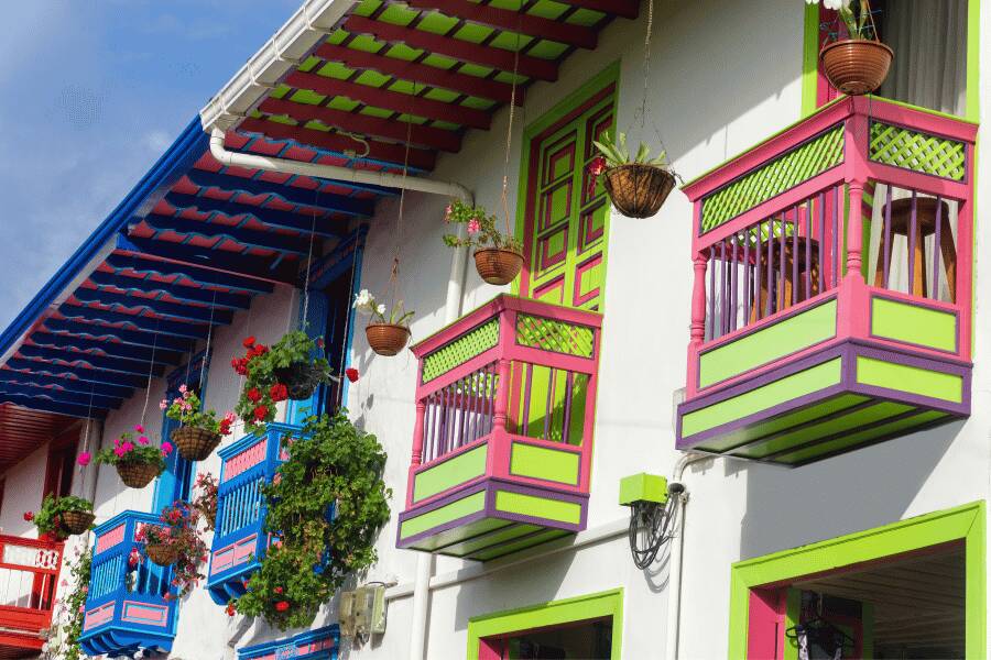 Des balcons en bois peints de couleurs vives, vert, rose et bleu, ornés de jardinières suspendues fleuries, décorent la façade blanche d'un bâtiment colonial sous un avant-toit coloré.