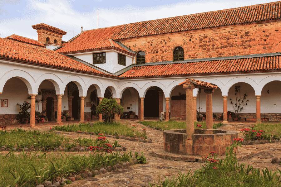 Un puits en pierre s'élève au centre d'une cour pavée et herbeuse, entourée par les arcades blanches et les toits en tuiles romaines d'un bâtiment colonial.