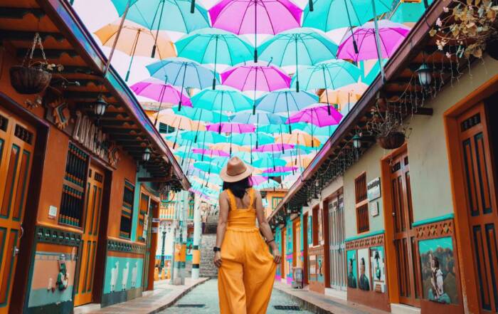Une femme en combinaison orange et chapeau marche de dos dans une rue pavée étroite, bordée de façades colorées et surmontée d'un toit de parapluies multicolores suspendus.