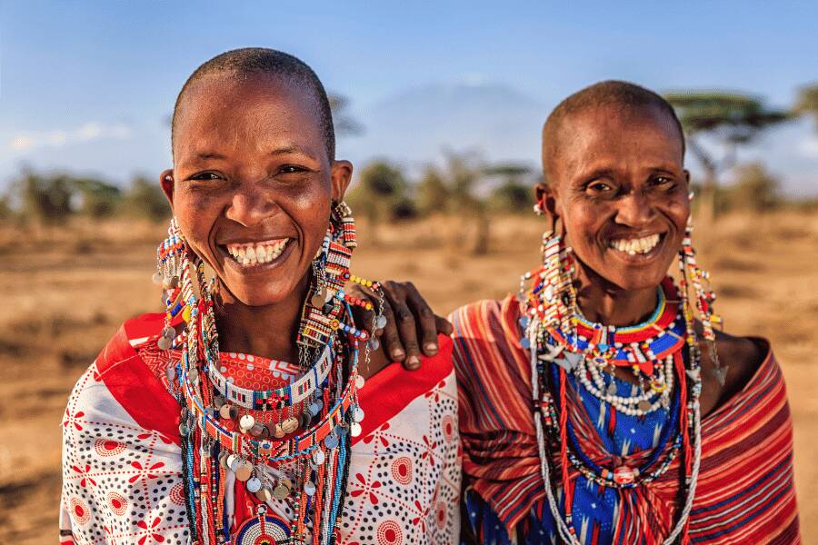 Deux femmes souriantes vêtues de tissus colorés et de bijoux en perles élaborés se tiennent côte à côte dans une plaine aride et ensoleillée.