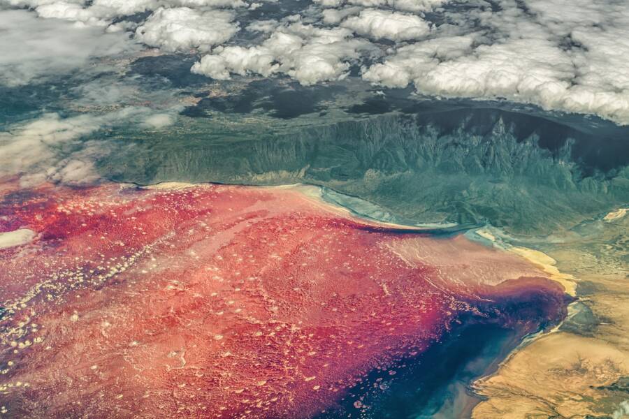 Une vue aérienne révèle les eaux teintées de rouge et de rose d'un grand lac côtoyant un paysage montagneux verdoyant sous un ciel nuageux.