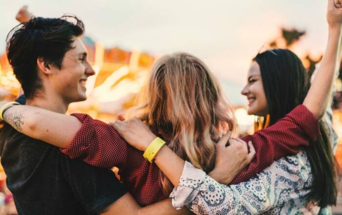 Trois jeunes amis s'enlacent joyeusement par les épaules, vus de dos, face au flou lumineux des manèges et attractions d'une fête foraine au soleil couchant.