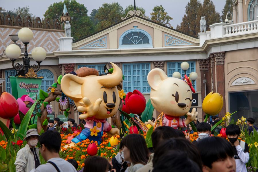 Deux statues géantes de mascottes souriantes accueillent les visiteurs au milieu d'un parterre de tulipes colorées, devant des bâtiments de style classique.
