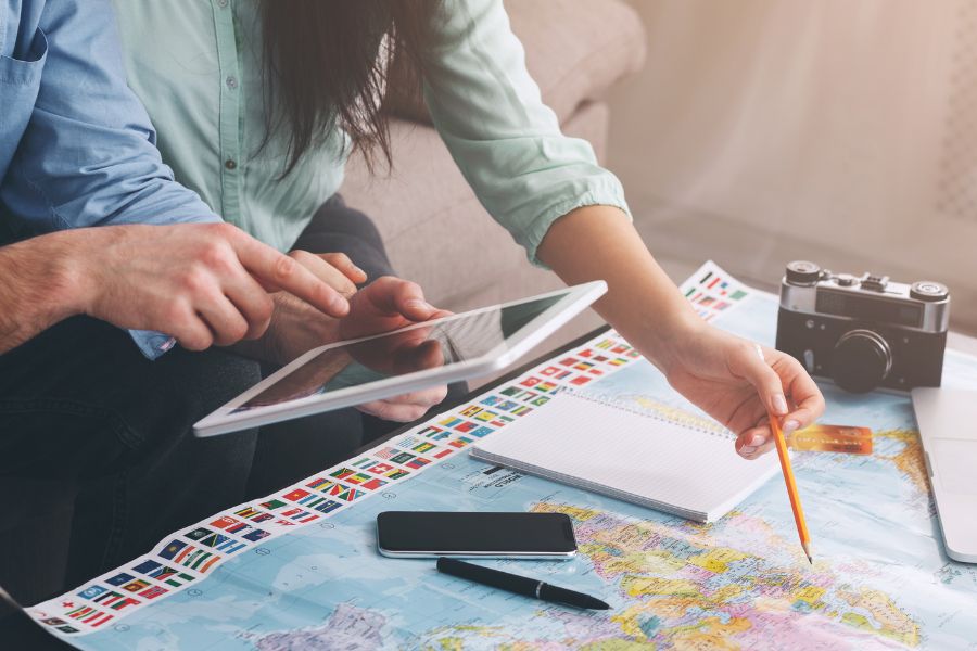 Une personne pointe du doigt une carte routière étalée sur une table en bois à côté d’un ordinateur portable, d’une boussole et d’accessoires.