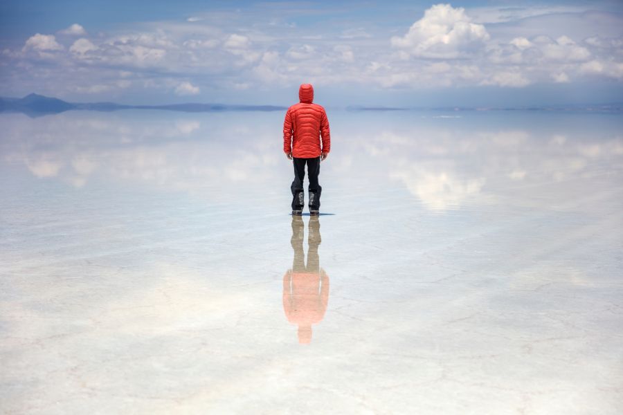 Une personne vêtue d'une doudoune rouge se tient de dos sur une étendue d'eau calme reflétant le ciel nuageux comme un miroir.