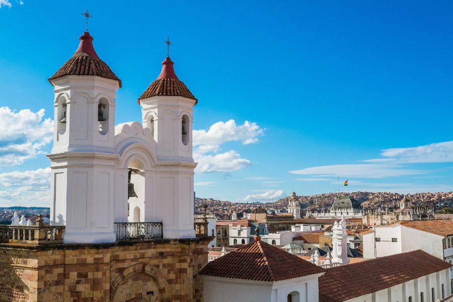 Deux clochers blancs aux toits de tuiles rouges s'élèvent au-dessus d'un panorama de bâtiments coloniaux sous un ciel bleu parsemé de nuages.