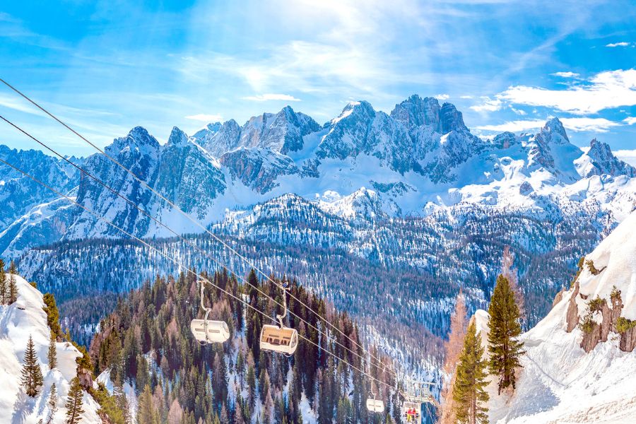 Des télésièges montent au-dessus d'une forêt enneigée face aux imposants sommets rocheux des Dolomites baignés de soleil.