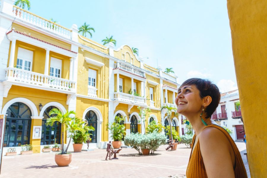 Une femme souriante regarde un grand bâtiment colonial jaune aux arcades et balcons blancs depuis une cour ombragée sous un ciel bleu.