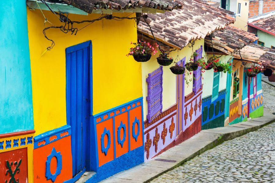 Des maisons traditionnelles aux façades peintes de couleurs vives et ornées de suspensions florales bordent une rue pavée en pente.
