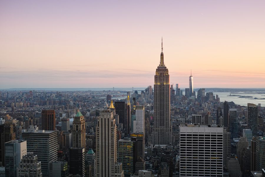 L'Empire State Building domine le panorama urbain de Manhattan au coucher du soleil, entouré de nombreux gratte-ciels sous un ciel aux teintes rosées.
