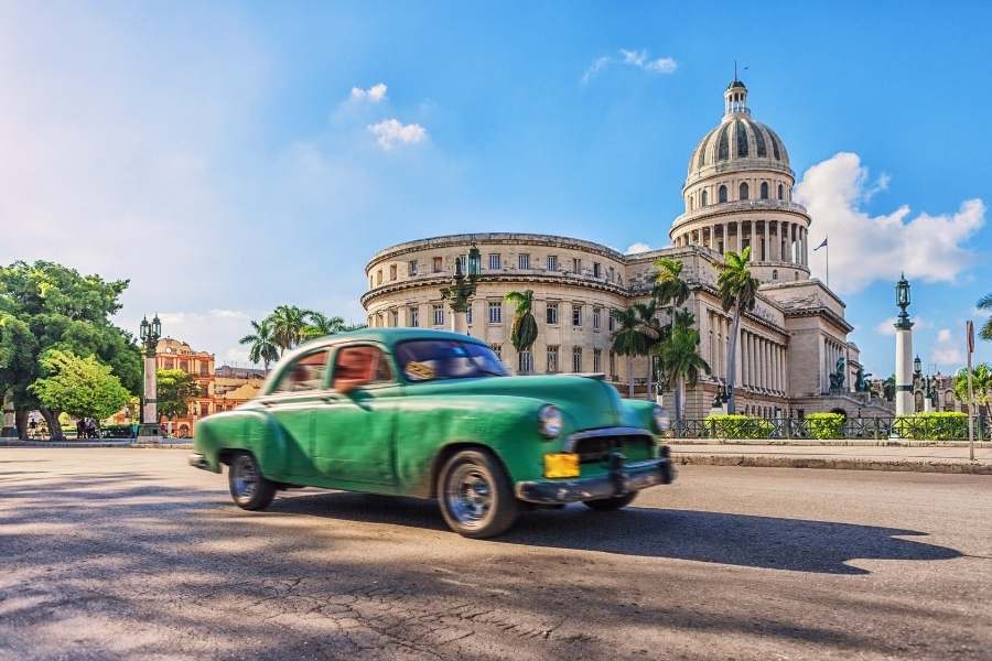 Une voiture ancienne verte roule sur une large avenue pavée devant un imposant capitole blanc surmonté d'un dôme, sous un ciel bleu parsemé de nuages.