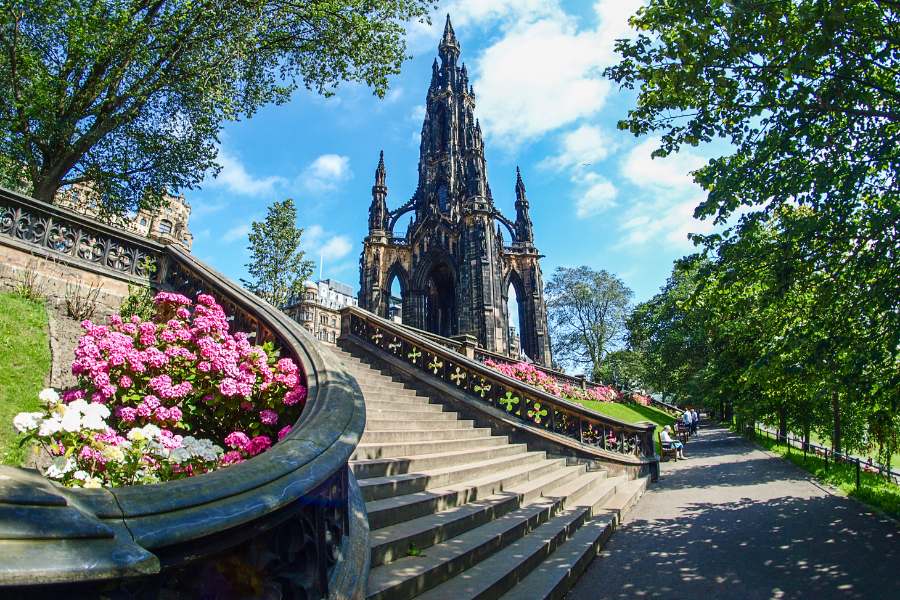 Un monument gothique en flèche noire s'élève majestueusement au-dessus d'un escalier de pierre orné de fleurs roses, bordant une allée ombragée dans un parc.