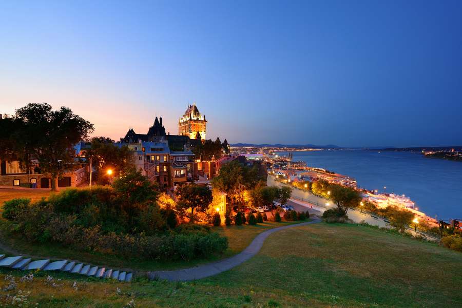Un grand château illuminé domine un fleuve et un quartier historique en contrebas, photographié depuis un parc herbeux incluant un sentier au crépuscule.