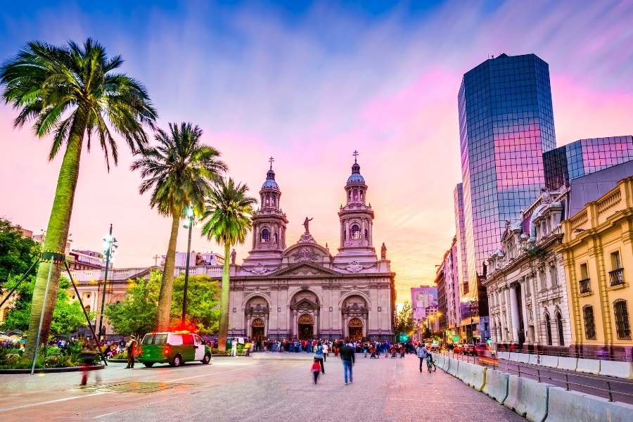 Des piétons marchent sur une place urbaine menant à une cathédrale historique entourée de gratte-ciels modernes et de grands palmiers, sous un ciel coloré de rose.