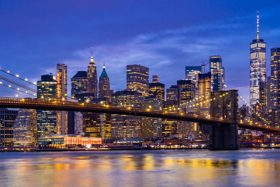 Un grand pont suspendu en pierre et acier enjambe un fleuve urbain devant les gratte-ciels illuminés de la ville à la tombée de la nuit.