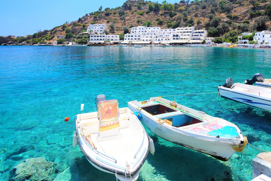 Des petits bateaux de pêche flottent sur une eau turquoise et transparente devant un village aux maisons blanches adossé à une colline rocheuse.
