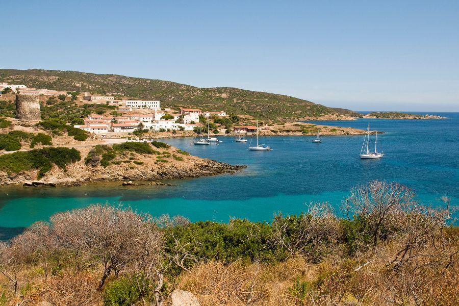 Des voiliers mouillent dans une baie aux eaux turquoise, bordée par une côte aride et des maisons blanches sous un ciel dégagé.