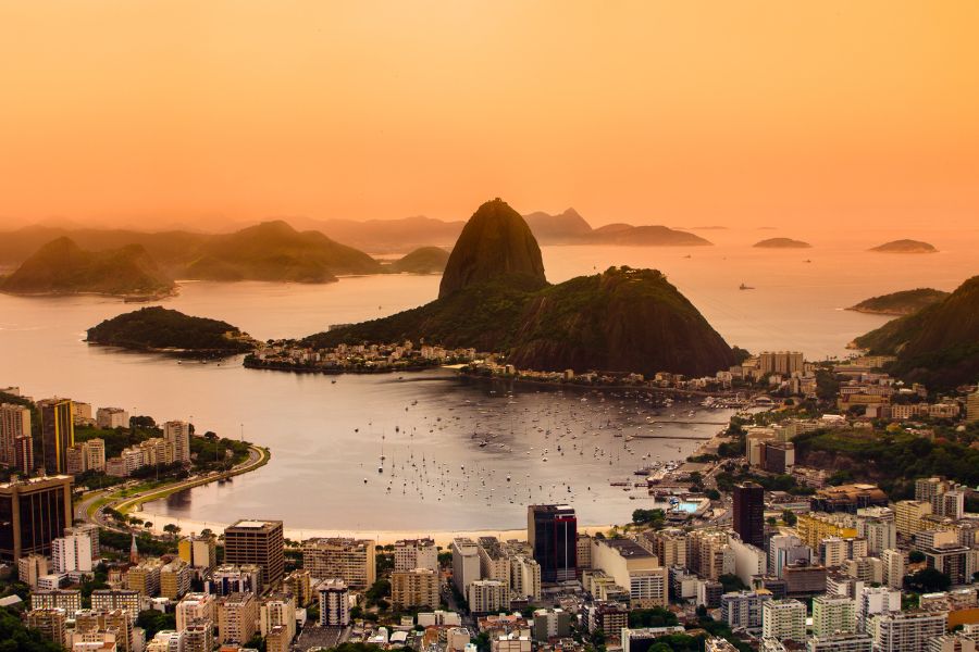 Une baie urbaine parsemée de voiliers s'étend devant une montagne escarpée au coucher du soleil, entourée de gratte-ciels sous une lumière dorée.
