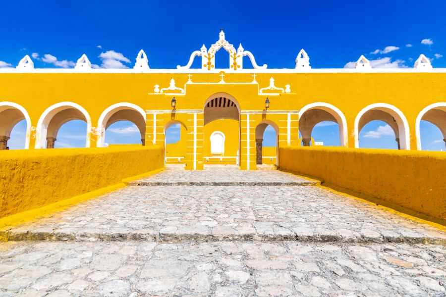 Une rampe pavée en pierre mène à une arche centrale au milieu d'une longue colonnade coloniale peinte en jaune vif et blanc sous un ciel bleu intense.
