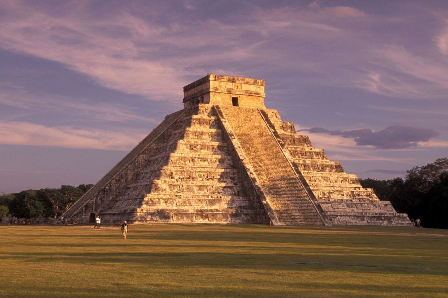 Une imposante pyramide maya en pierre s'élève au centre d'une vaste esplanade verdoyante sous un ciel bleu ensoleillé parsemé de quelques nuages.