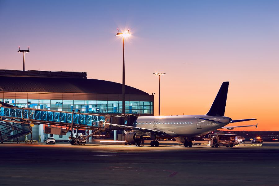 Un avion stationne devant un terminal d'aéroport au crépuscule, relié à une passerelle d'embarquement. Les lumières illuminent la scène, évoquant un départ calme et une atmosphère moderne, paisible nocturne accueillante.