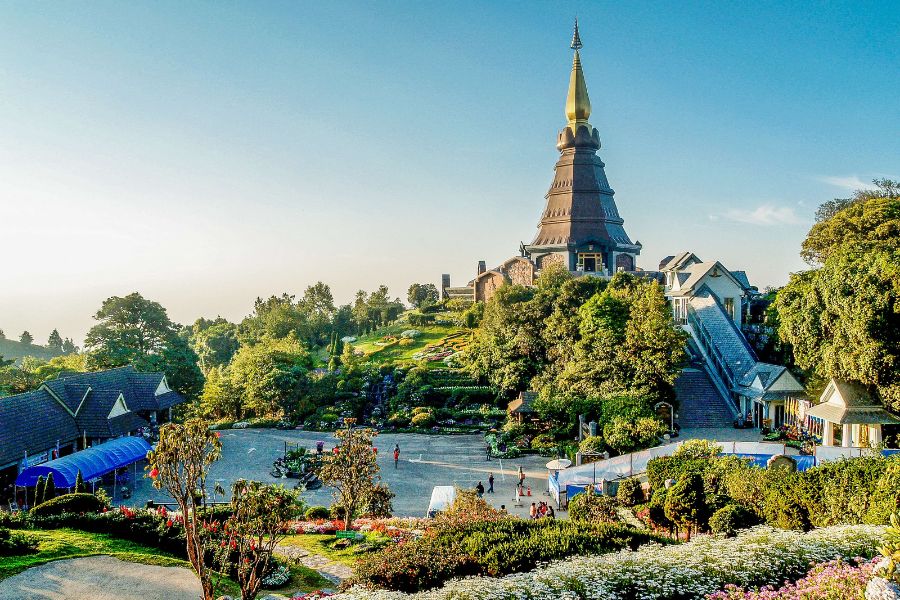Un stupa royal orné s'élève au sommet d'une montagne verdoyante bordée de jardins fleuris sous un ciel nuageux au coucher du soleil.