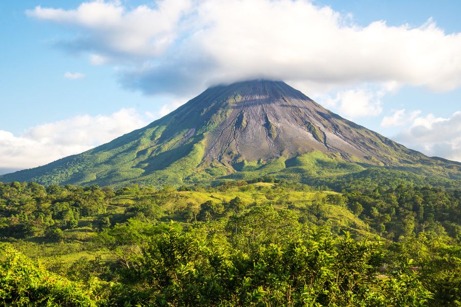 Un volcan au sommet conique s'élève au-dessus d'une forêt tropicale dense et verdoyante sous un ciel bleu parsemé de nuages.