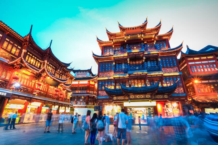 Quartier traditionnel chinois animé au crépuscule, avec bâtiments illuminés aux architectures anciennes, foule en mouvement dans la rue piétonne et ambiance urbaine chaleureuse mêlant culture, lumière et modernité asiatique vibrante.