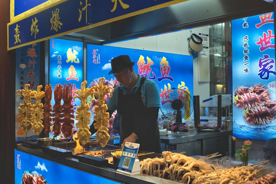 Vendeur de rue chinois préparant des brochettes et fruits de mer grillés dans un stand éclairé, entouré d’affiches colorées et d’aliments typiques, créant une atmosphère nocturne authentique et gourmande.