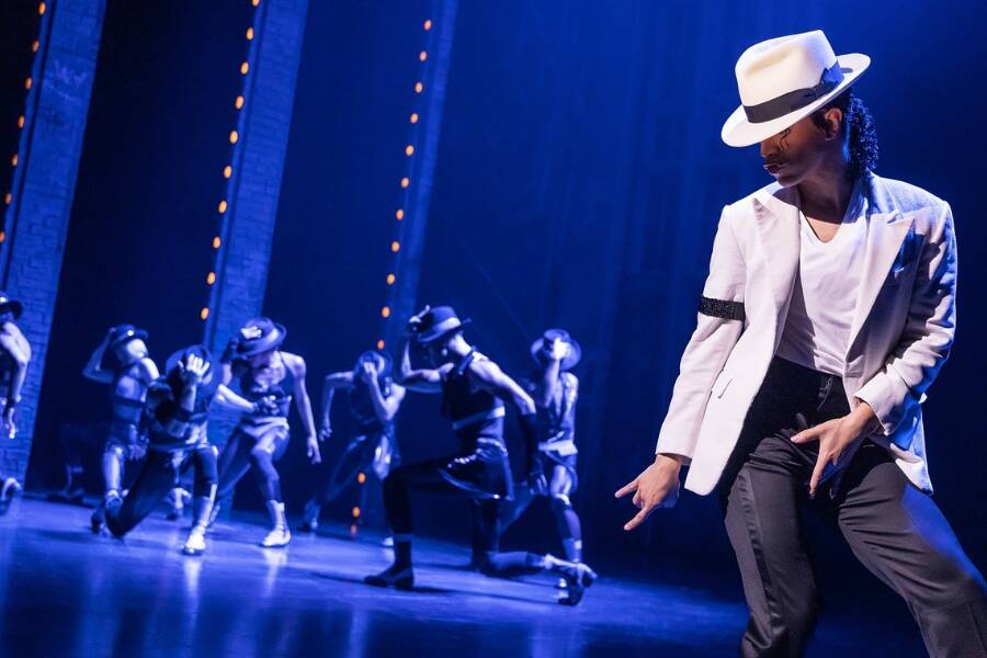 Un danseur en costume blanc et chapeau en feutre assorti exécute un pas de danse iconique au premier plan, tandis qu'une troupe de danseurs en arrière-plan reproduit sa posture sous des projecteurs bleus.