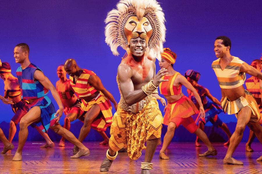Un danseur portant une coiffe de lion mène une chorégraphie dynamique, entouré d'autres artistes en tenues colorées aux motifs africains sur une scène illuminée de bleu.