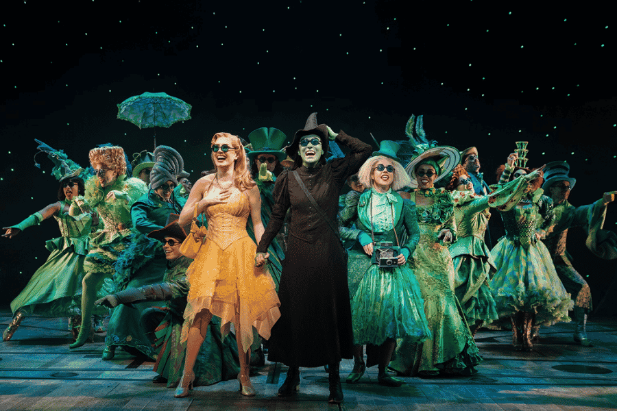 Une troupe de comédiens vêtus de costumes extravagants verts et jaunes, portant des lunettes de soleil noires, pose joyeusement sur scène sous un fond constellé de lumières.