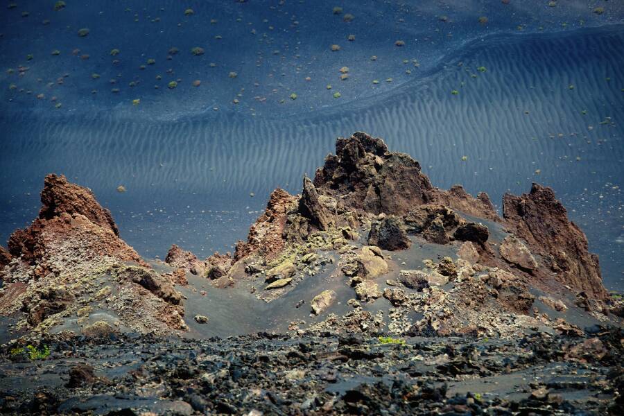 Une crête rocheuse volcanique et aride du parc national de Timanfaya surplombe un vaste désert de sable noir marqué de ridules, parsemé de quelques petites touffes de végétation éparses.