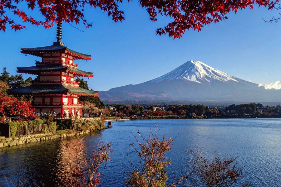 Une pagode traditionnelle rouge et blanche se dresse au bord d'un lac paisible, face au mont Fuji enneigé, sous un ciel bleu encadré par des feuilles d'érable rouges.