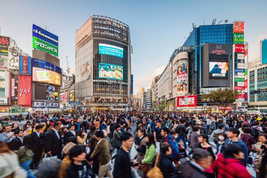 Une foule compacte de piétons traverse un grand carrefour urbain entouré de gratte-ciels modernes recouverts d'immenses écrans publicitaires lumineux et d'enseignes colorées en japonais et en anglais.