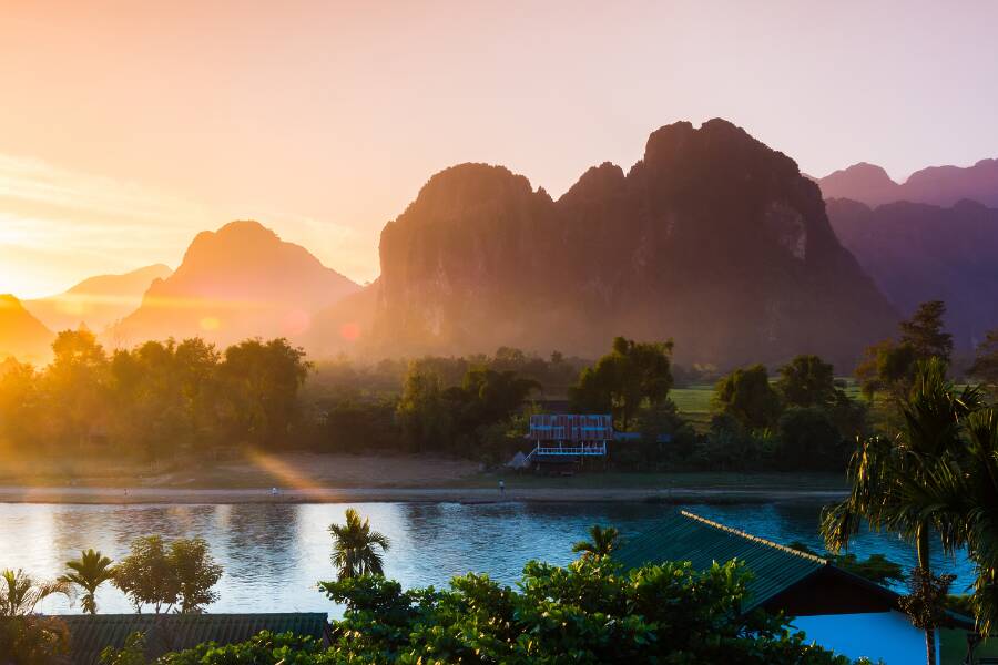 Une rivière calme coule devant un village verdoyant dominé par de hautes montagnes au coucher du soleil, illustrant le Laos comme un joyau méconnu de l'Asie.