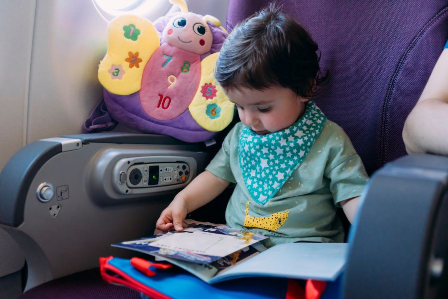 Un jeune enfant assis sur un siège d'avion feuillette un livret, à côté d'un coussin en forme de papillon coloré posé contre l'accoudoir.