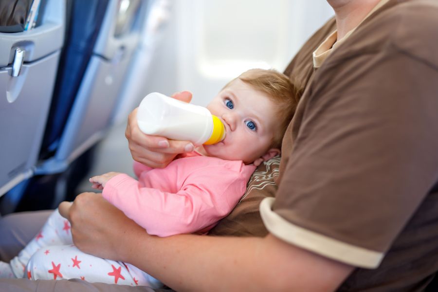 Un bébé aux yeux bleus boit son biberon, confortablement installé dans les bras d'un adulte assis sur un siège à bord d'un avion de ligne.