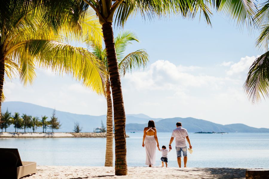 Un couple tenant leur enfant par la main marche sur une plage de sable blanc bordée de palmiers, face à une mer calme et des montagnes lointaines.