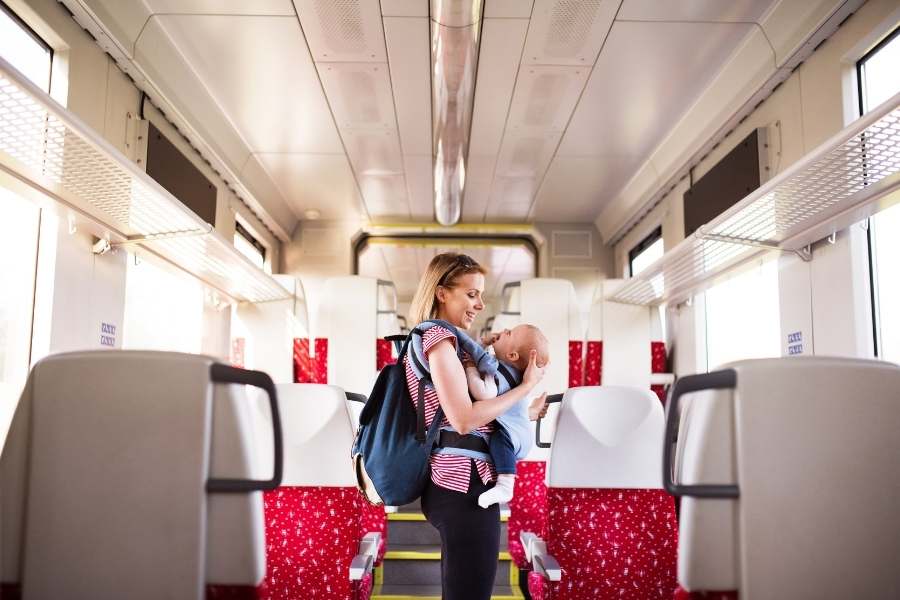 Une femme souriante portant son sac à dos et son bébé dans un porte-bébé ventral se tient debout au milieu de l'allée centrale d'un train.