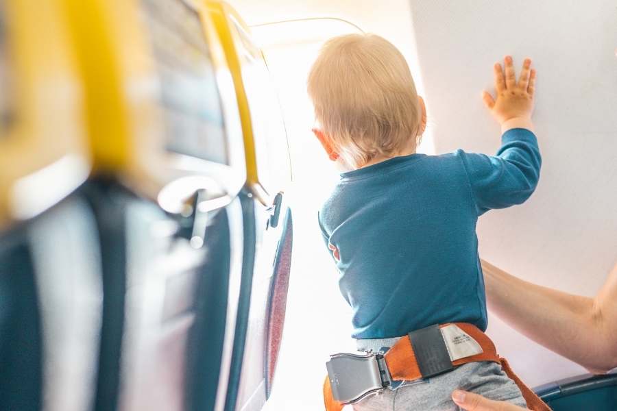 Un jeune enfant équipé d'une ceinture de sécurité orange se tient debout face à la paroi intérieure lumineuse d'un avion, soutenu par une main adulte.