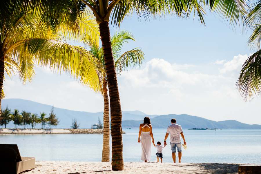 Un couple tenant leur enfant par la main marche sur une plage de sable blanc bordée de palmiers, face à une mer calme et des montagnes lointaines.