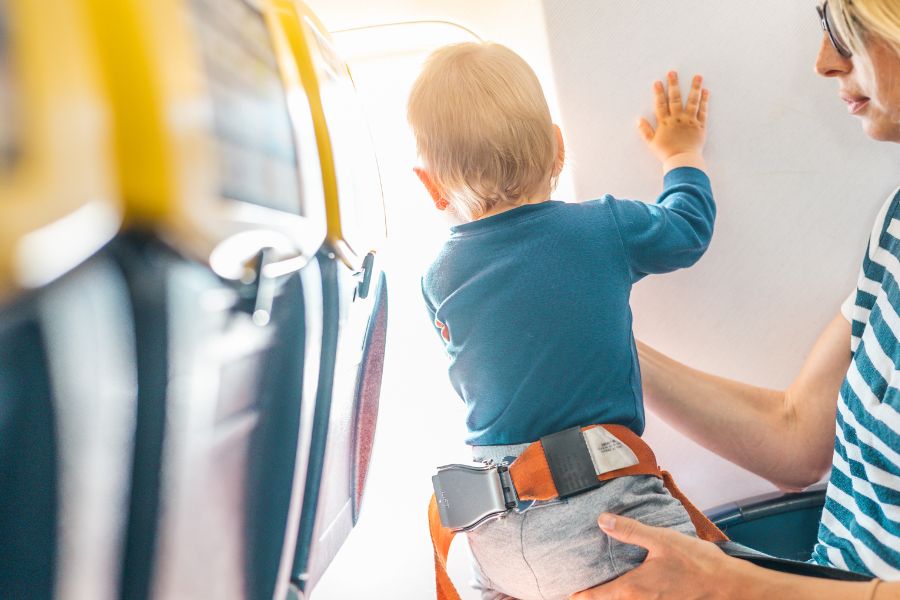 Un jeune enfant équipé d'une ceinture de sécurité orange se tient debout face à la paroi intérieure lumineuse d'un avion, soutenu par une main adulte.