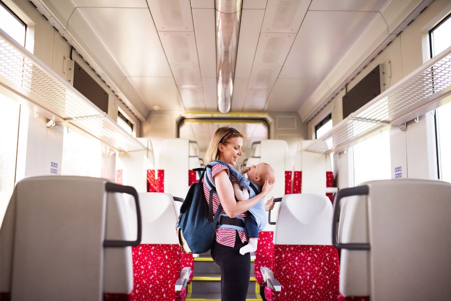 Une femme souriante portant son sac à dos et son bébé dans un porte-bébé ventral se tient debout au milieu de l'allée centrale d'un train.
