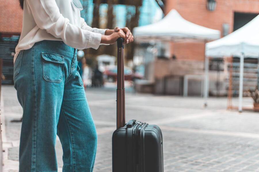 Une personne debout tient la poignée d’une valise noire dans une rue pavée. Elle paraît attendre un transport, prête à partir pour un voyage ou revenir.