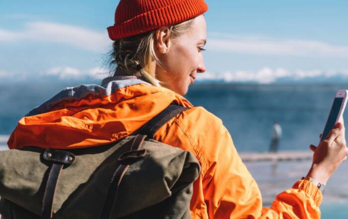 Une voyageuse, portant un bonnet rouge, une veste orange et un sac à dos, consulte son téléphone face à la mer. Elle semble préparer son itinéraire ou vérifier des informations utiles.