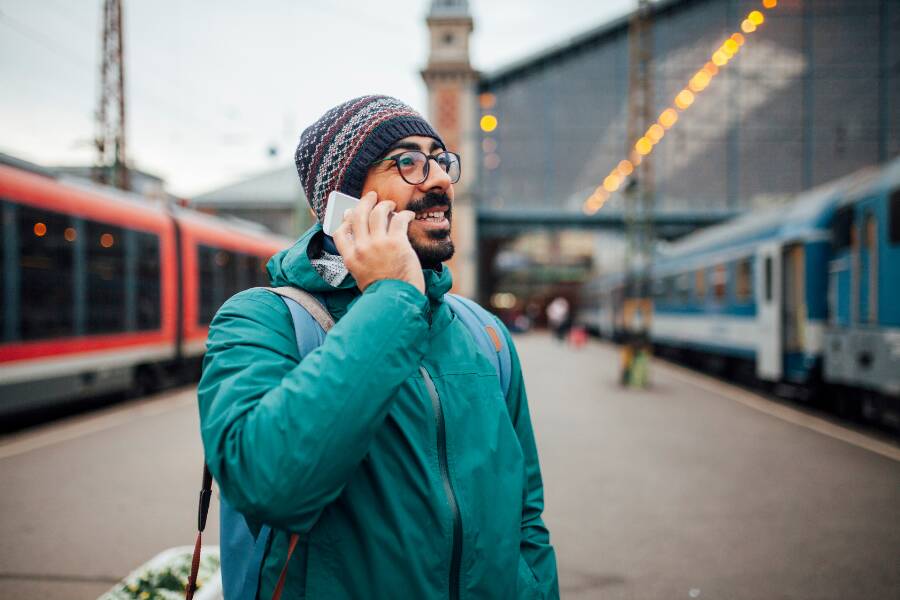 Un voyageur, habillé chaudement avec un bonnet et une veste verte, téléphone sur un quai de gare. Il paraît organiser son trajet ou demander des renseignements utiles.