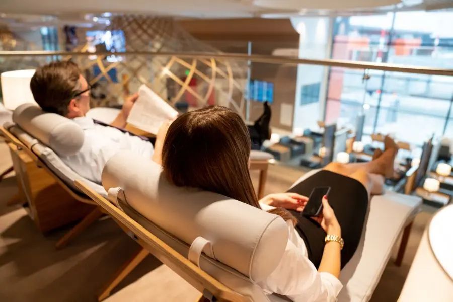 Des voyageurs se détendent confortablement dans un salon VIP d’aéroport, profitant d’un espace calme avec fauteuils inclinables, ambiance élégante, vue sur le terminal et moment de repos avant leur vol.