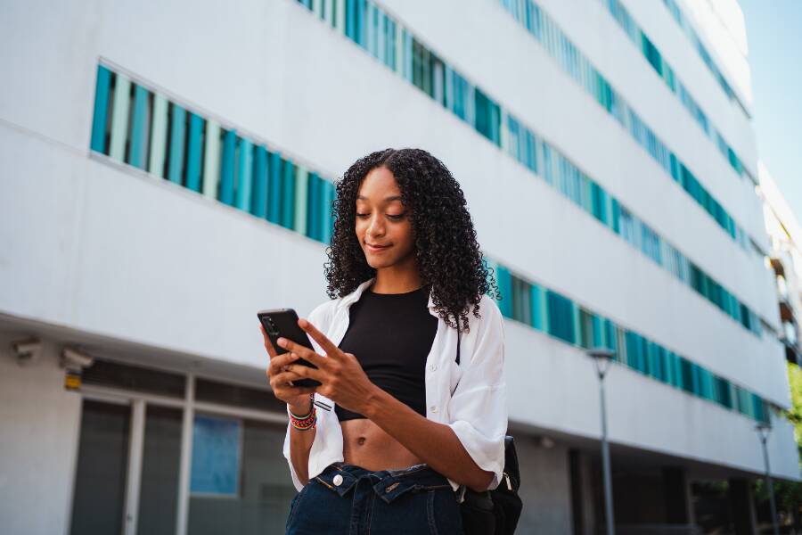 Une jeune femme consulte son smartphone devant un bâtiment moderne. Elle semble lire un message, utiliser une application de voyage ou rechercher des informations pratiques avant son déplacement.